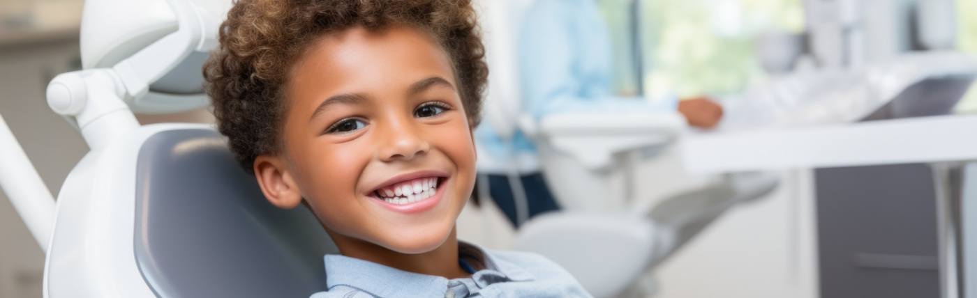 A little boy seated in a dentist’s chair and smiling while waiting to see the children’s dentist