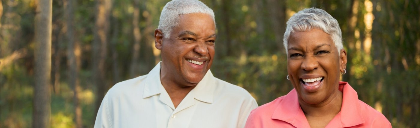 Older man and woman smiling outdoors with trees in background
